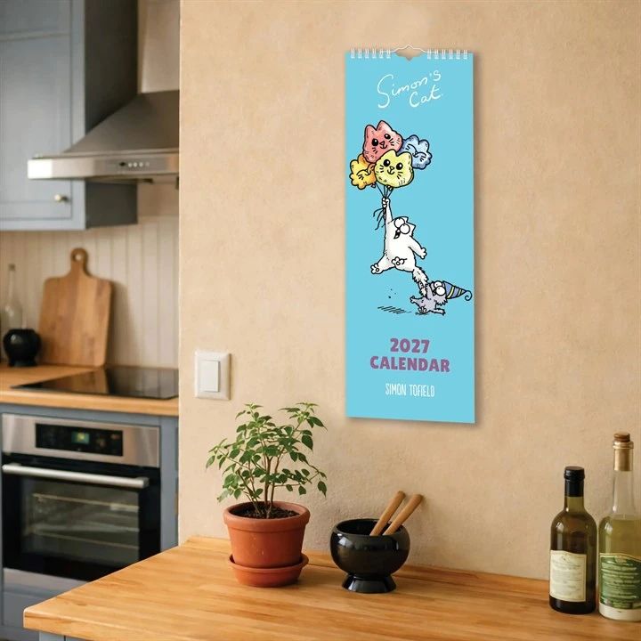 A 2027 Simon Tofield calendar featuring a cartoon cat with balloons, hanging on a beige kitchen wall above a wooden countertop 