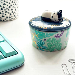 Decorative container with a black and white cat figurine on top, surrounded by office supplies on a light surface.
