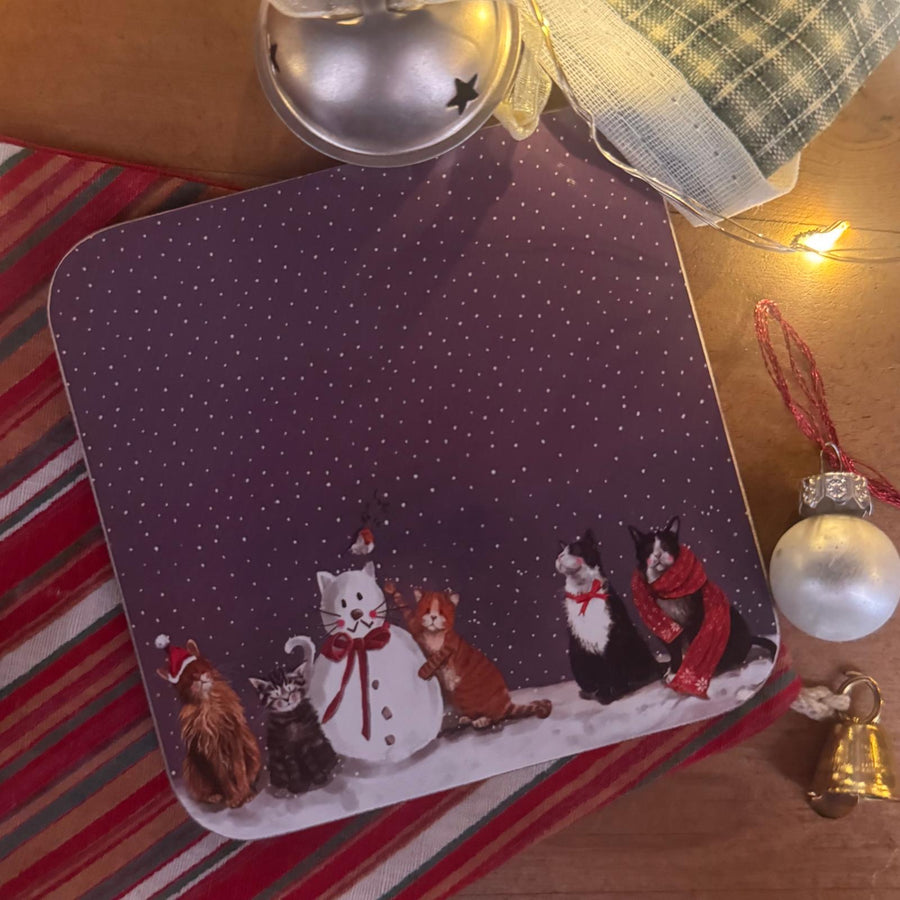 A coaster featuring a winter scene with several cats in hats and scarves on a snowy ground under a dark purple starry sky. 