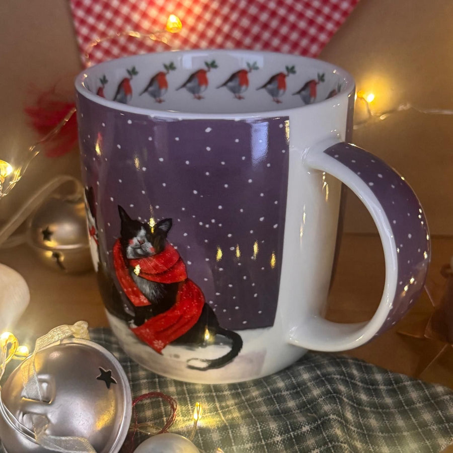 A ceramic mug with a purple side panel featuring a black cat in a red scarf under falling snow