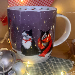 A festive ceramic mug with a purple winter scene featuring two black and white cats in snow, one wearing a red scarf