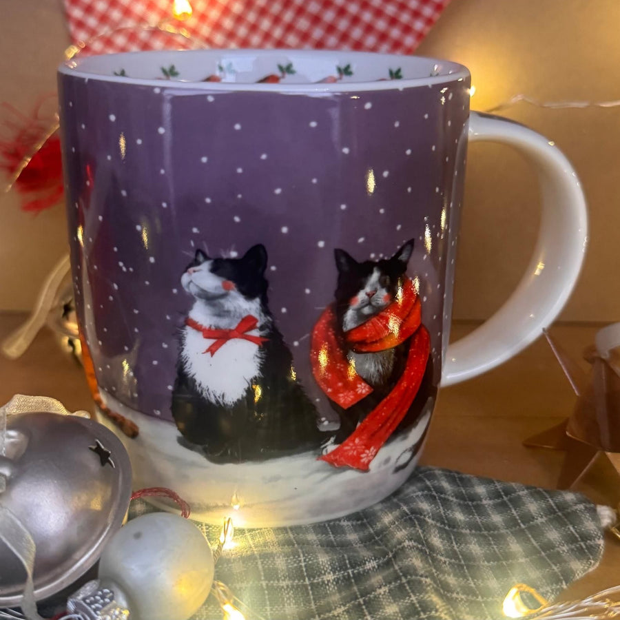 A festive ceramic mug with a purple winter scene featuring two black and white cats in snow, one wearing a red scarf