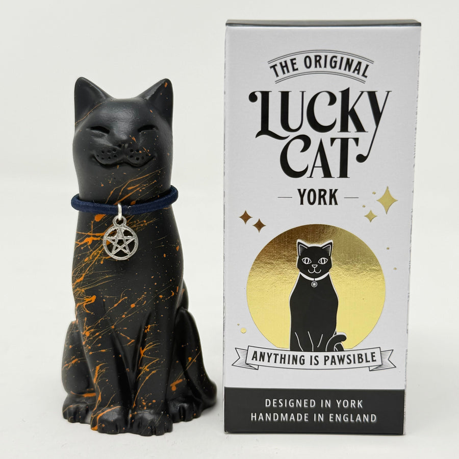 A black cat figurine with orange paint splatters and a pewter pentagram pendant sits next to its white and gold branded packaging box labeled 'The Original Lucky Cat York'.