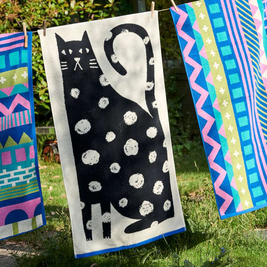 Patterned towels hanging on a clothes line outdoors, with a white spotted black cat print towel in the centre