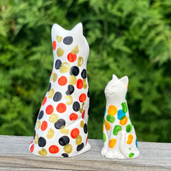 Two resin cat figurines with colourful polka dot patterns on a wooden surface with a green blurred background.