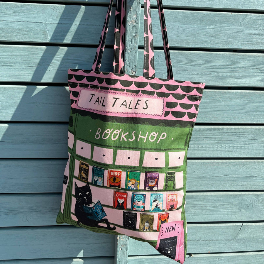 A fabric tote bag with a bookshop theme, featuring a green and pink design with a black cat and book illustrations