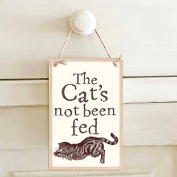 A rectangular hanging wall sign with a cream background, reading “The Cat’s not been fed” above a brown illustration of a cat