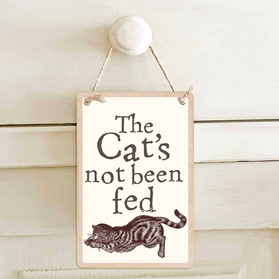 A rectangular hanging wall sign with a cream background, reading “The Cat’s not been fed” above a brown illustration of a cat