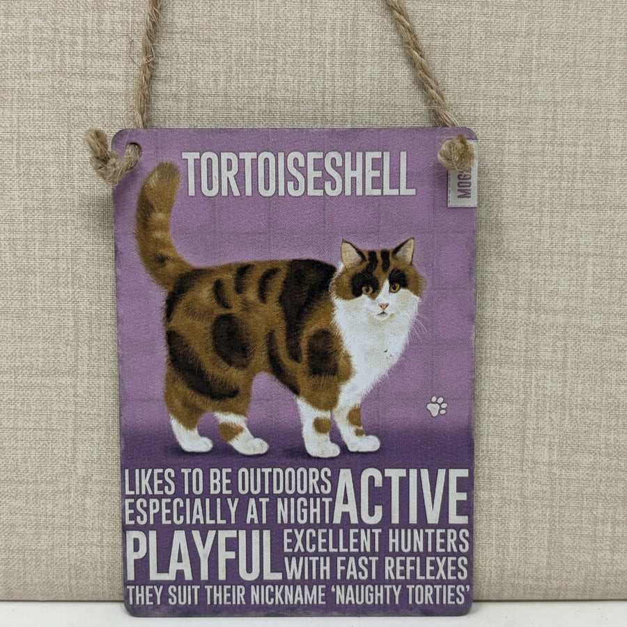 A rectangular hanging sign with a purple background featuring an illustration of a tortoiseshell cat and text that reads 'TORTOISESHELL' and additional cat-related phrases.