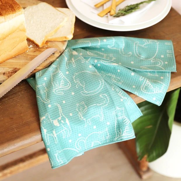 A turquoise kitchen towel with a white cat pattern draped over a wooden table next to sliced bread and a plate.