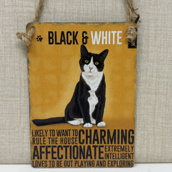 A rustic hanging sign, featuring a black and white cat illustration. The sign reads 'BLACK & WHITE' at the top and humorous cat traits at the bottom, hanging by twine