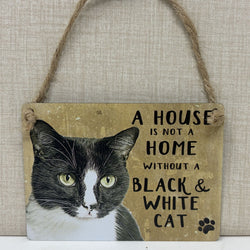 A rustic hanging sign with a black-and-white cat portrait and the text 'A HOUSE IS NOT A HOME WITHOUT A BLACK & WHITE CAT' 