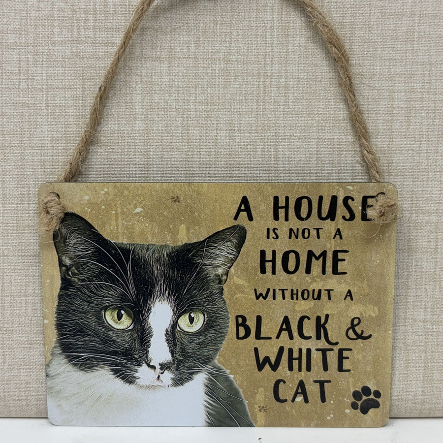 A rustic hanging sign with a black-and-white cat portrait and the text 'A HOUSE IS NOT A HOME WITHOUT A BLACK & WHITE CAT' 