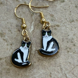 A pair of gold-tone dangle earrings shaped like black-and-white cats, photographed on a textured stone surface.