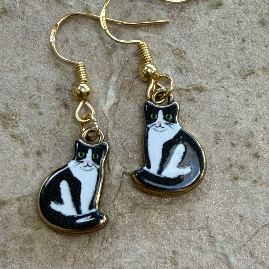 A pair of gold-tone dangle earrings shaped like black-and-white cats, photographed on a textured stone surface.