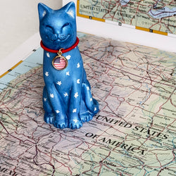 A blue ceramic cat figurine with white star patterns and a red collar featuring an American flag medallion, displayed on top of a United States map.