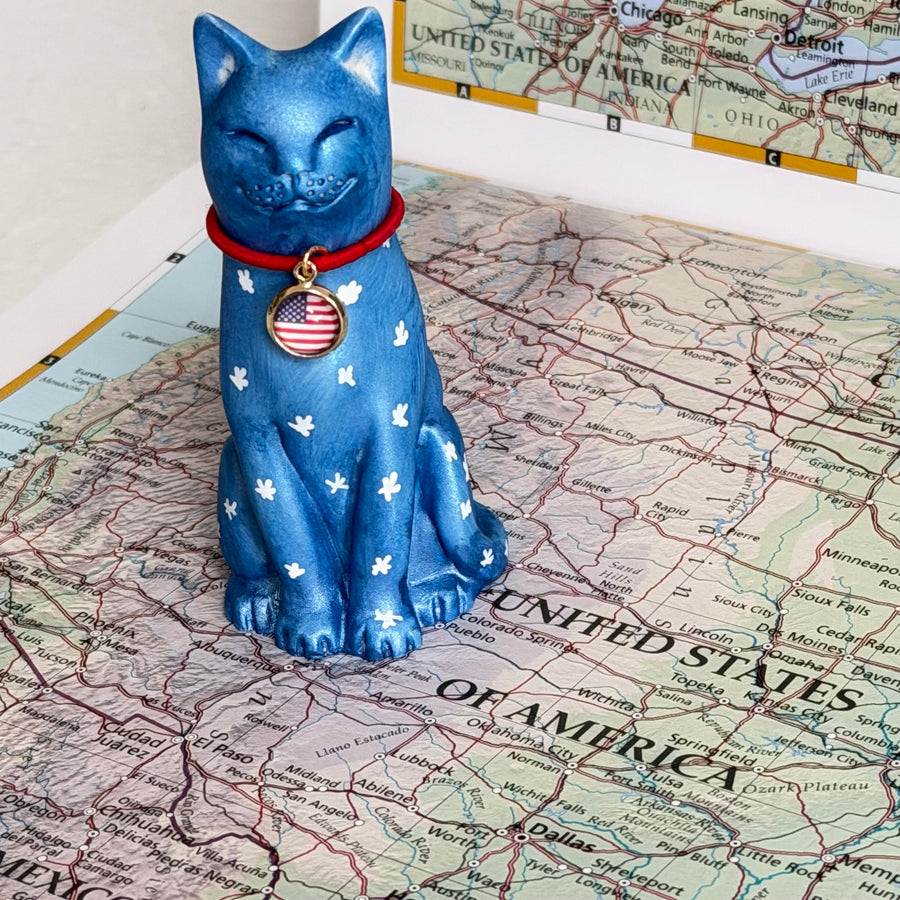 A blue ceramic cat figurine with white star patterns and a red collar featuring an American flag medallion, displayed on top of a United States map.