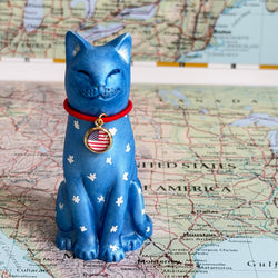 A blue cat figurine with white paw prints and a red collar featuring a US flag medallion, standing on a map background.