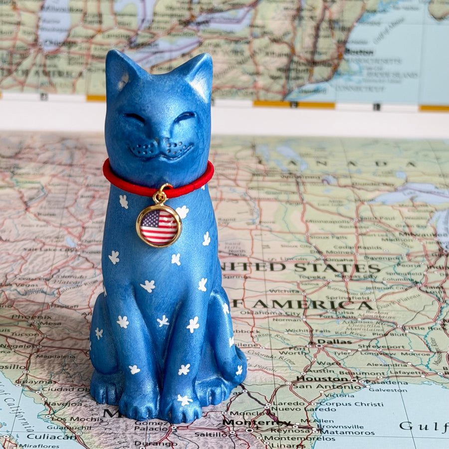 A blue cat figurine with white paw prints and a red collar featuring a US flag medallion, standing on a map background.