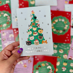 A hand with purple nail polish holds a light blue greeting card featuring a Christmas tree made of illustrated cat faces, with the text 'WE WISH YOU A MEOWY CHRISTMAS'. 