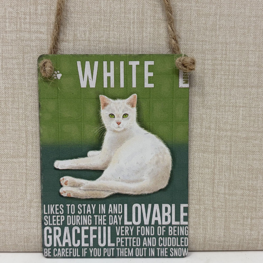 A rectangular hanging wall sign with a photo of a white cat on a green background and printed text, suspended by twine from a beige fabric wall.