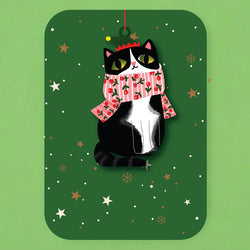 Black and white cat hanging wooden ornament attached to a greetings cardl wearing a pink stripy scarf on a green background with festive decorations.