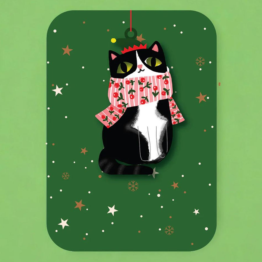 Black and white cat hanging wooden ornament attached to a greetings cardl wearing a pink stripy scarf on a green background with festive decorations.