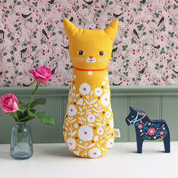 A yellow cat-shaped plush cushion with a white floral pattern and embroidered face details, styled on a white table with pink roses in a glass vase