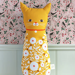 A yellow cat-shaped decorative pillow with a white floral print on the lower half, featuring embroidered facial details and a red collar, set against a pink floral wallpaper background.
