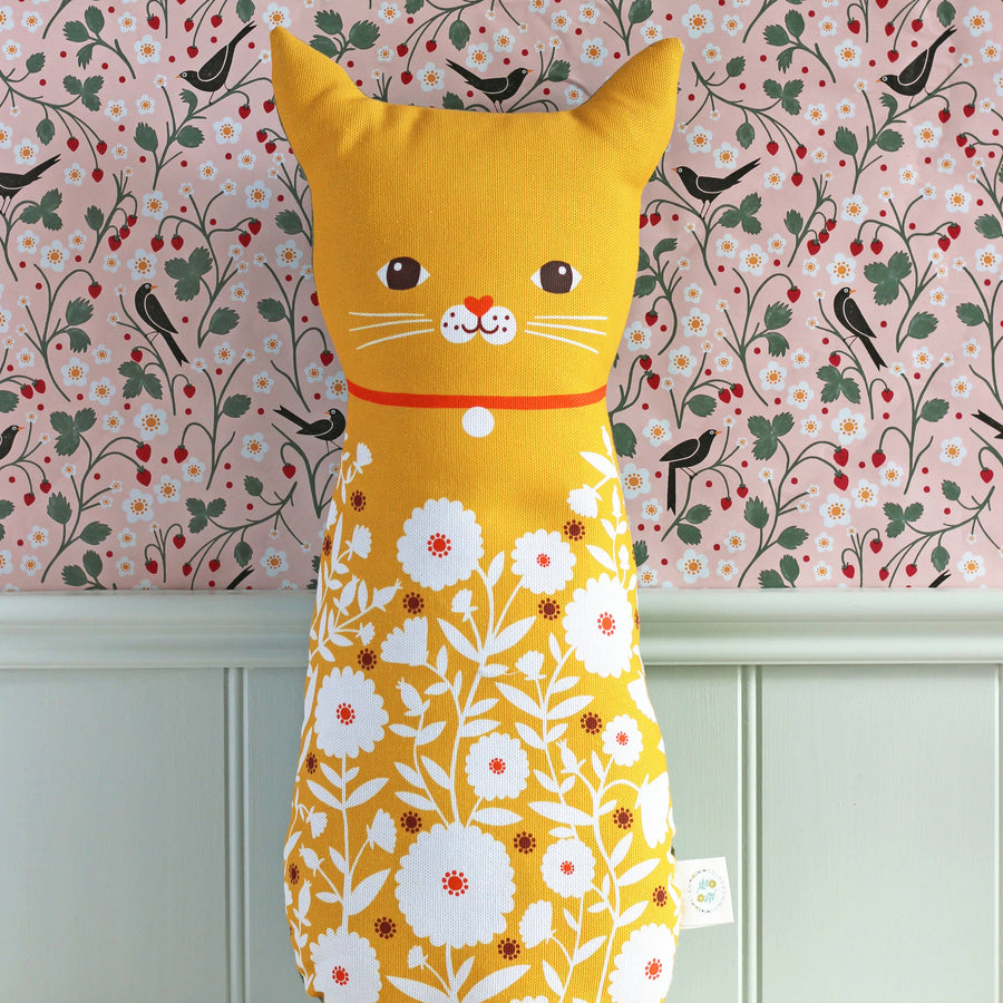 A yellow cat-shaped decorative pillow with a white floral print on the lower half, featuring embroidered facial details and a red collar, set against a pink floral wallpaper background.