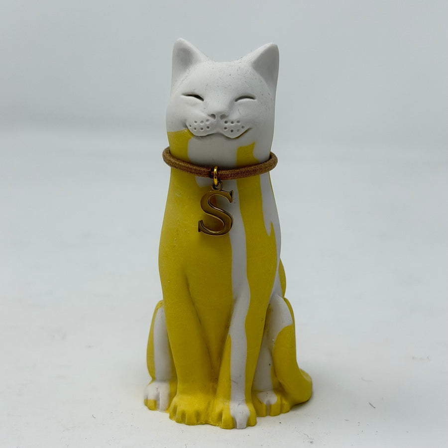 A resin lucky cat figurine in yellow and white, wearing a brown collar with a gold letter 'S' charm