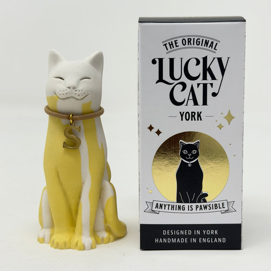 A white and yellow lucky cat figurine wearing a brown collar with a gold letter 'S' pendant, shown next to its white and gold retail box 'The Original Lucky Cat York'