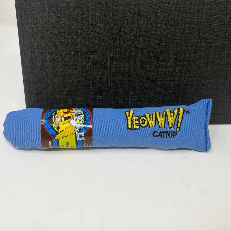 A blue fabric catnip toy with yellow embroidered 'Yeoww! Catnip' text, shown on a white surface with a dark textured box in the background.