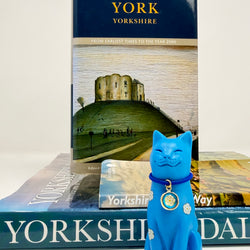 A blue cat ornament decorated with white roses with blue collar with round white rose charm, displayed with books about Yorkshire