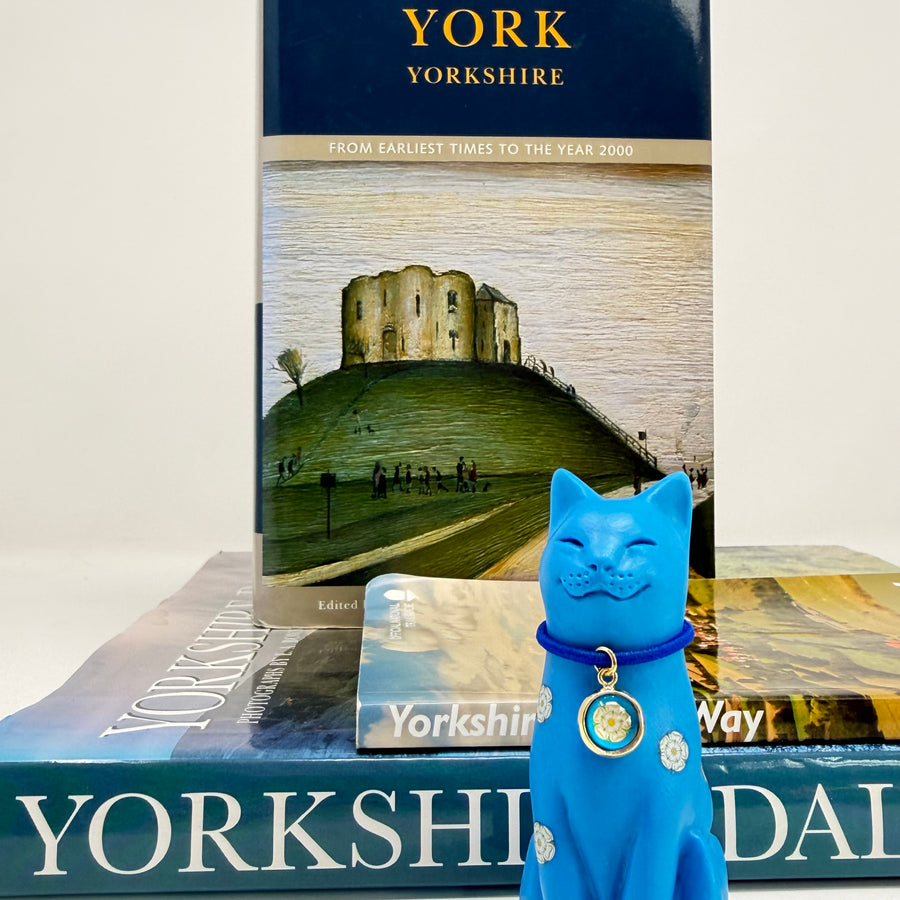 A blue cat ornament decorated with white roses with blue collar with round white rose charm, displayed with books about Yorkshire