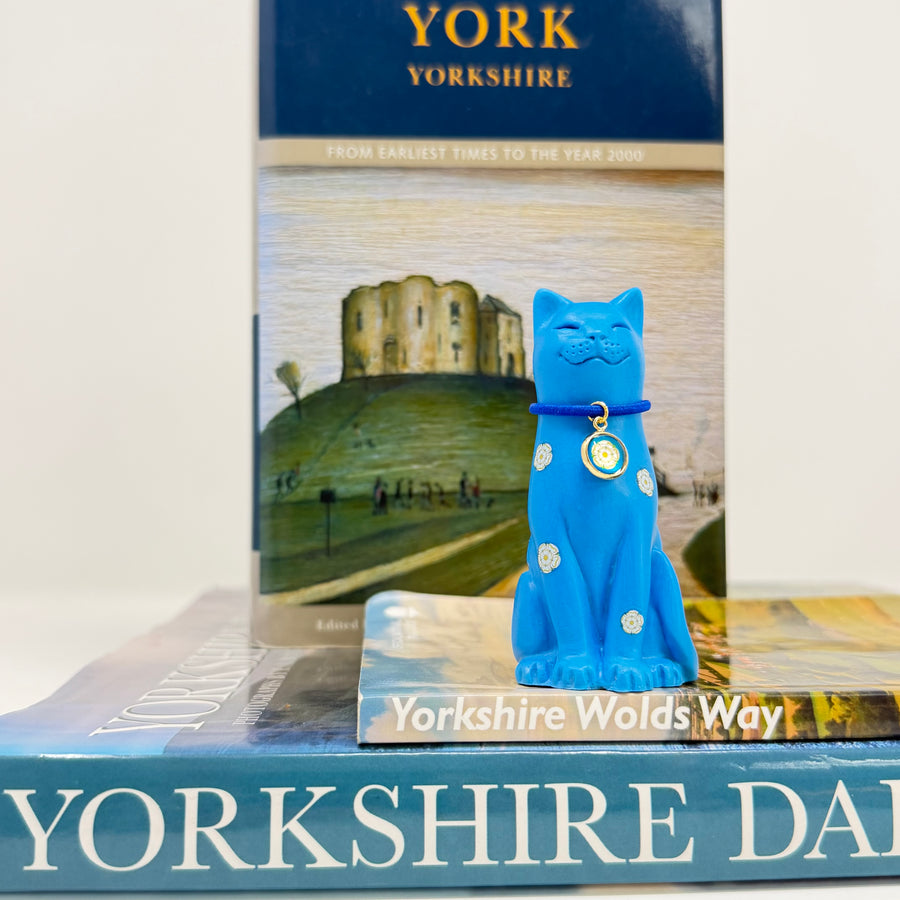 A blue cat ornament decorated with white roses with blue collar with round white rose charm, displayed with books about Yorkshire