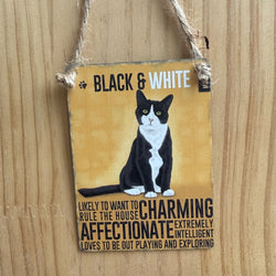 A rectangular metal hanging sign, with black & white cat illustration and the words 'BLACK & WHITE' at the top. The sign includes text describing alleged attributes of black & white cats and is suspended by twine against a wooden wall.