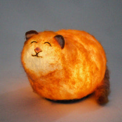 A hand-felted, ginger cat shaped LED night light emitting a soft glow, with a smiling face and black ears.