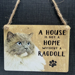 A rustic rectangular metal hanging sign with a ragdoll cat illustration and the phrase 'A HOUSE IS NOT A HOME WITHOUT A RAGDOLL' in black text, plus a black paw print. The sign hangs from twine against a dark textured background.