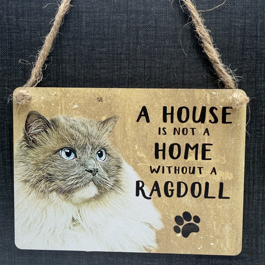 A rustic rectangular metal hanging sign with a ragdoll cat illustration and the phrase 'A HOUSE IS NOT A HOME WITHOUT A RAGDOLL' in black text, plus a black paw print. The sign hangs from twine against a dark textured background.