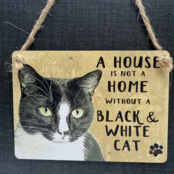 A rustic hanging metal sign with a black-and-white cat illustration and the phrase 'A HOUSE IS NOT A HOME WITHOUT A BLACK & WHITE CAT' hanging from a twine loop