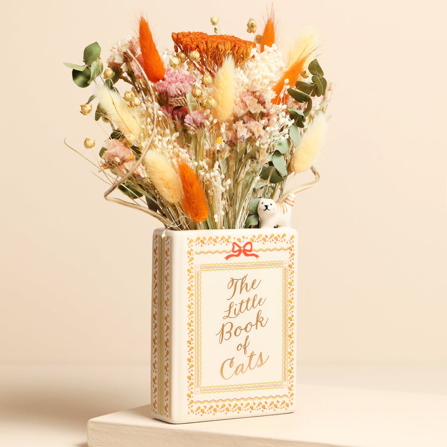 A decorative vase shaped like a book titled "The Little Book of Cats," filled with dried flowers and grasses, set against a warm beige background.