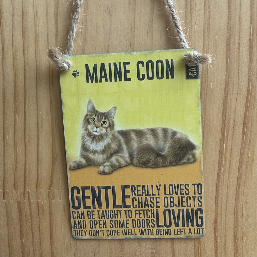 A rectangular metal hanging sign, with maine coon cat illustration and the word 'MAINE COON' at the top. The sign includes text describing alleged attributes of maine coon cats and is suspended by twine against a wooden wall.