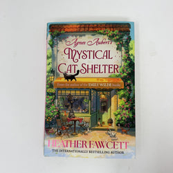The book cover of 'Agnes Aubert's Mystical Cat Shelter' by Heather Fawcett, featuring a whimsical illustration of a cat shelter with cats and flowers, photographed on a white background.