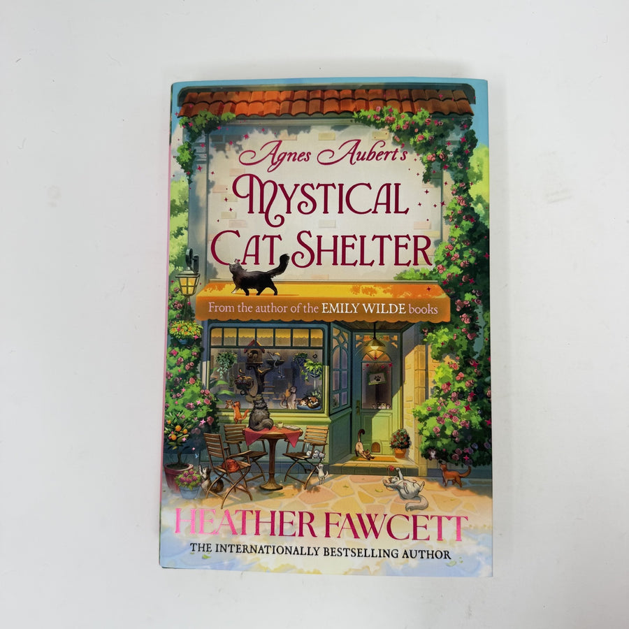 The book cover of 'Agnes Aubert's Mystical Cat Shelter' by Heather Fawcett, featuring a whimsical illustration of a cat shelter with cats and flowers, photographed on a white background.