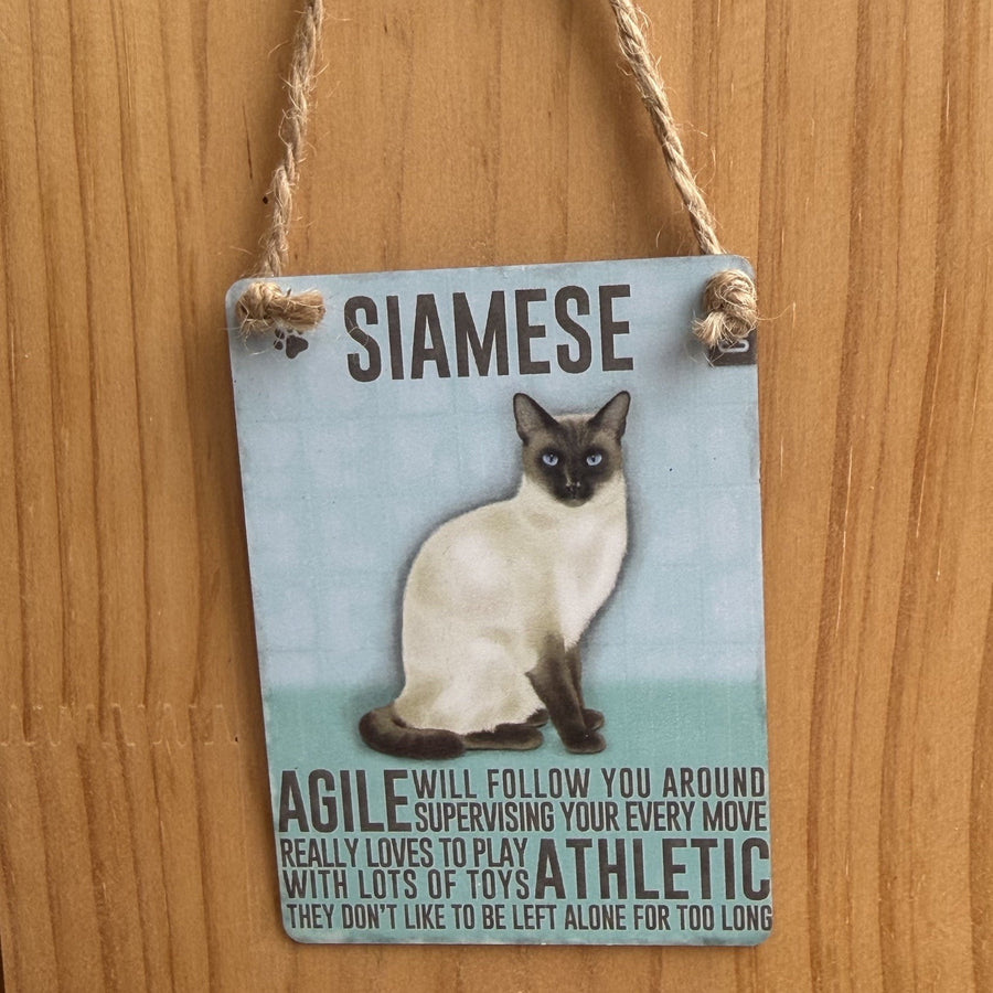 A rectangular metal hanging sign, with siamese cat illustration and the word 'SIAMESE' at the top. The sign includes text describing alleged attributes of  siamese cats and is suspended by twine against a wooden wall.