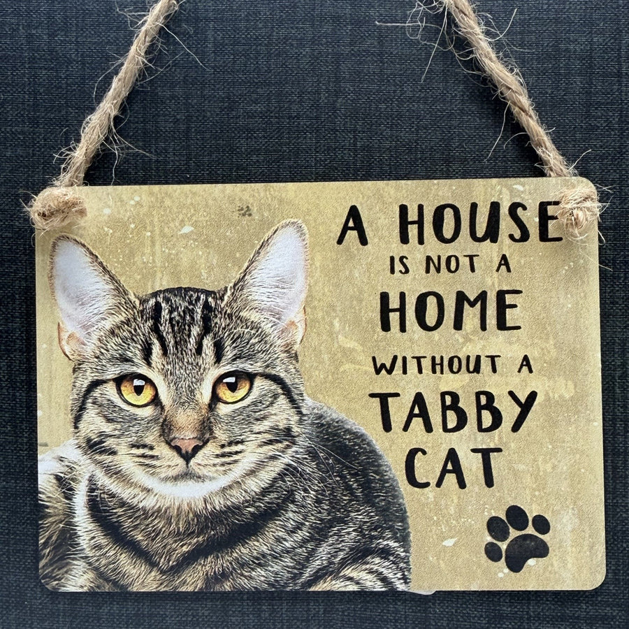 A rectangular hanging metal sign with a tabby cat and 'A HOUSE IS NOT A HOME WITHOUT A TABBY CAT' plus a black paw print, suspended by twine against a dark textured background.