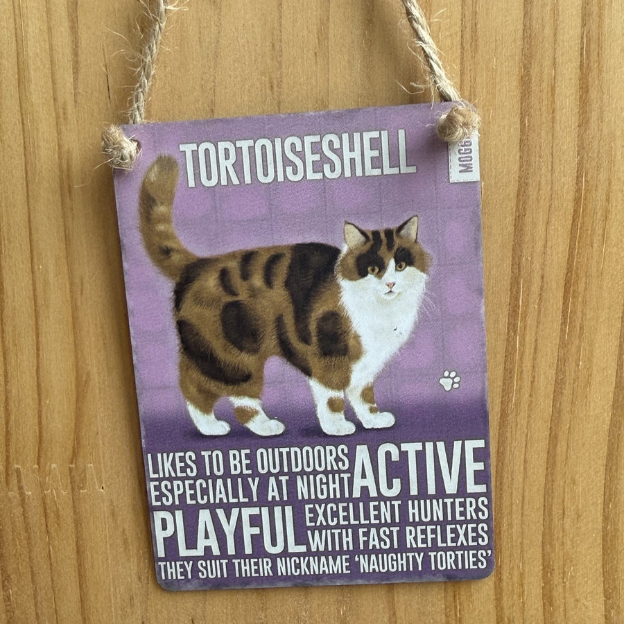 A rectangular metal hanging sign, with tortoiseshell cat illustration and the word 'TORTOISESHELL' at the top. The sign includes text describing alleged attributes of tortoiseshell cats and is suspended by twine against a wooden wall.